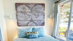 Un dormitori amb un llit, un mural amb un mapa del món, una finestra amb cortines, i un coixí amb la inscripció 'TRAILS LAKE'.