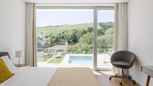 A bedroom with a large bed, a chair, and a view of a vineyard and swimming pool.
