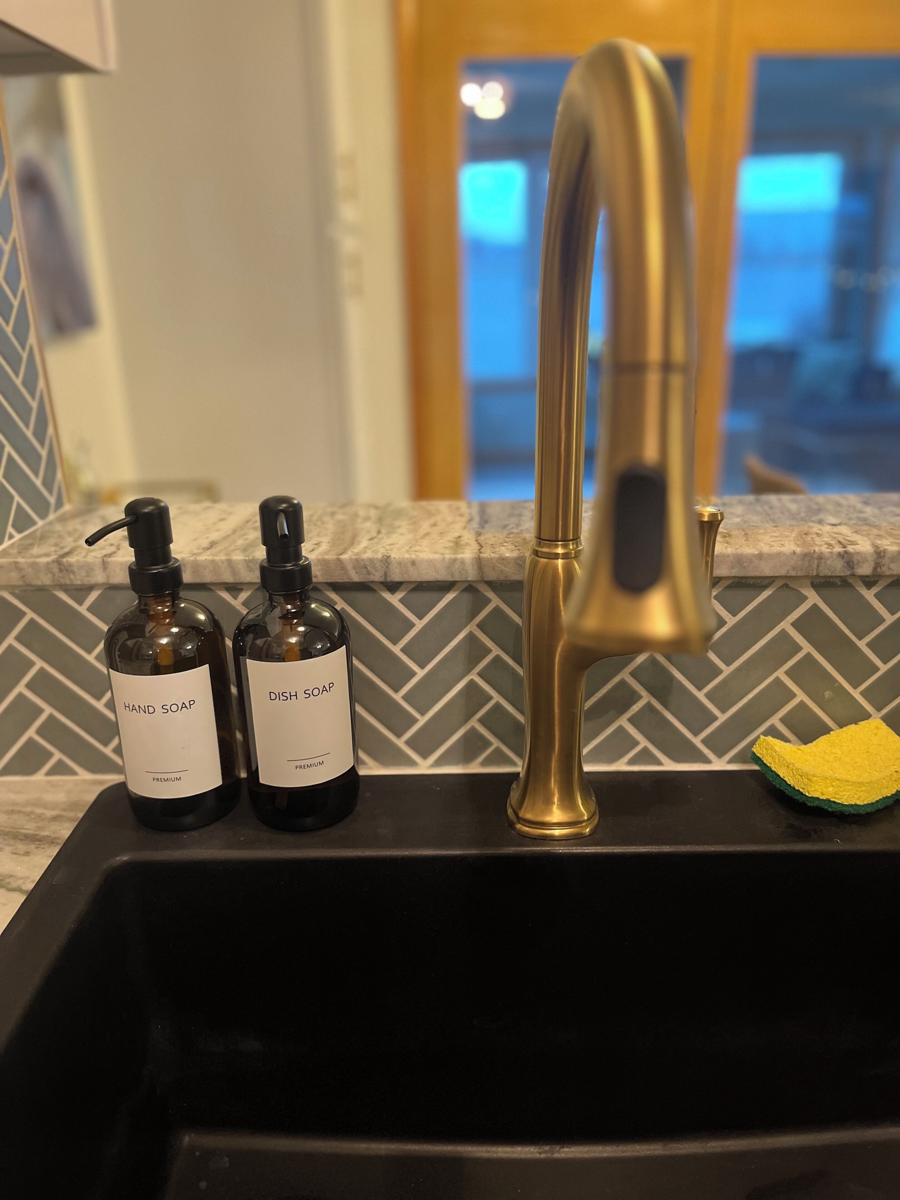 Ein Küchenspülbecken mit zwei Handseifenflaschen, beschriftet mit 'HAND SOAP' und 'DISH SOAP', einem goldfarbenen Wasserhahn und einem gelben Schwamm auf der Arbeitsfläche.