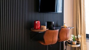 A modern hotel room with a flat-screen TV mounted on the wall, a small table with a coffee maker, two leather chairs, and a wicker side table with a potted plant. - Creative Corner: Stylish Apt. in Ghent's Heart (Gent)
