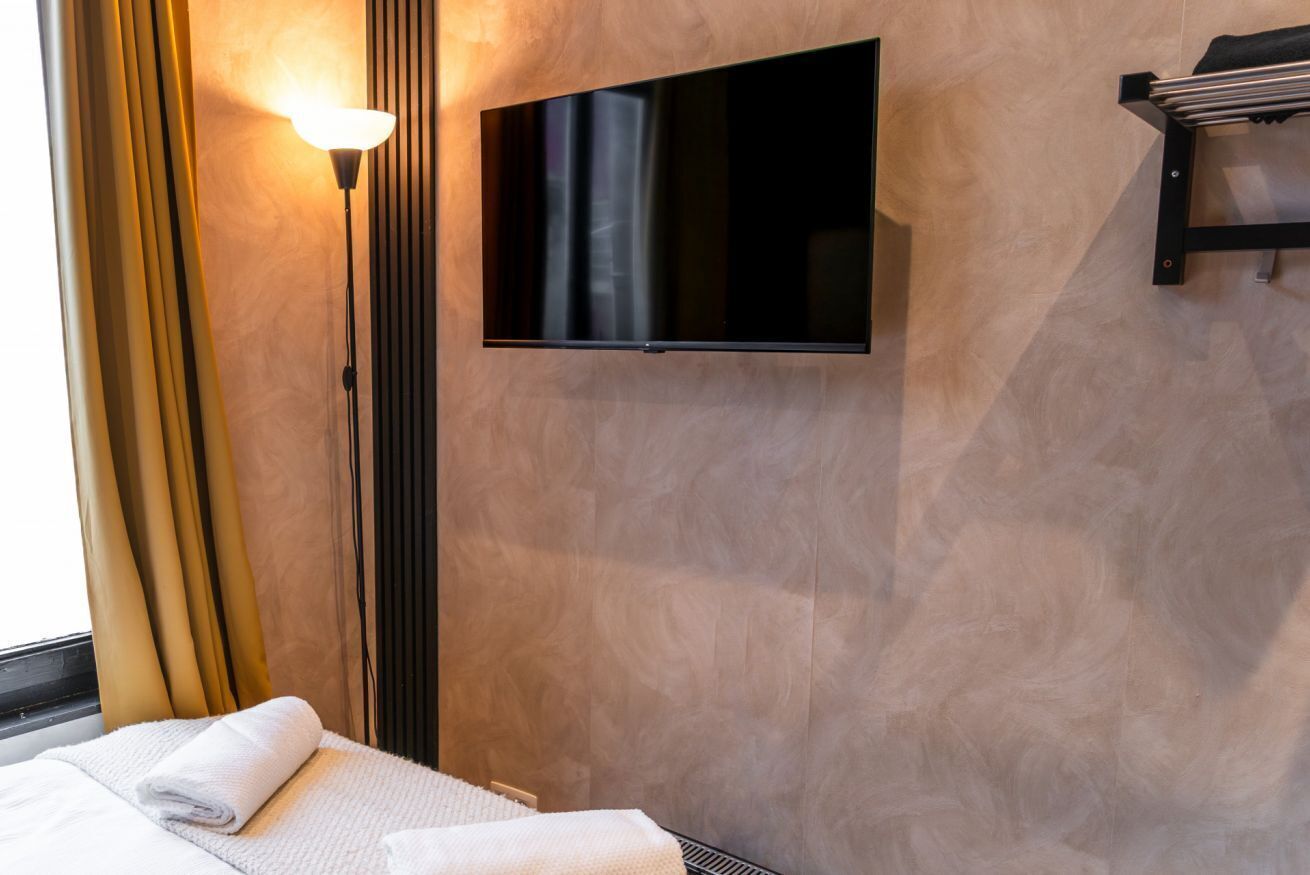 Une chambre d’hôtel équipée d’un téléviseur à écran plat fixé au mur, d’une lampe sur pied, d’un lit avec une serviette blanche et d’une fenêtre avec des rideaux.