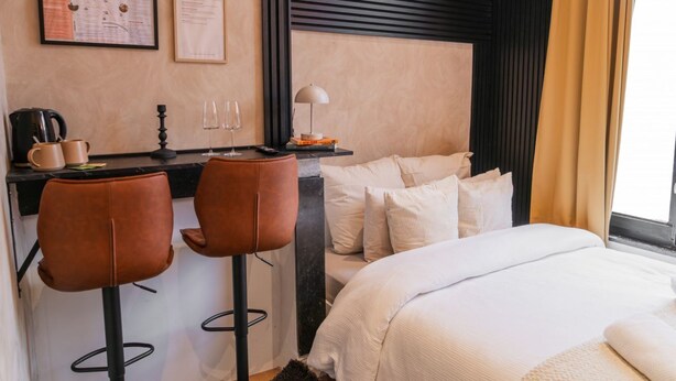 A hotel room with a bed, two bar stools, a desk with a coffee machine, a lamp, and a map on the wall.