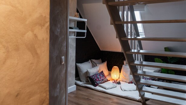 A loft bedroom with a wooden staircase, a cozy reading nook with a lamp, and a view of the outdoors.