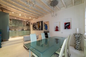 A modern interior with a glass dining table, white chairs, and a kitchen in the background.