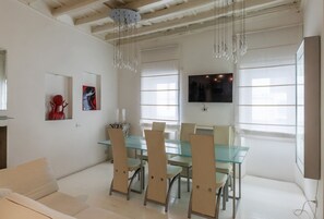 Un comedor moderno con una mesa y sillas de vidrio, un televisor montado en la pared y una araña.