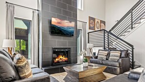A modern living room with a fireplace, grey sofas, a coffee table, and a staircase. - Serenity Now - Brand New Property/View/Indoor Pool/Hot Tub/Movie Room/Game Room (Sevierville)