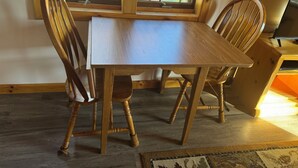 A wooden dining table with four chairs, a television on a stand, and a window with a view of a wooden deck and greenery. - Cozy 2 bedroom cottage with full kitchen minutes from Pictured Rocks Cruises! (Munising)