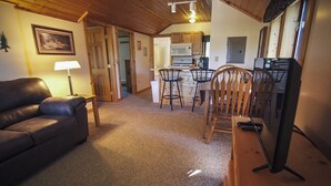A cozy living area with a leather sofa, a wooden dining table with chairs, a TV, and a kitchen in the background. - Cozy 2 bedroom cottage with full kitchen minutes from Pictured Rocks Cruises! (Munising)