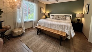 Quarto com cama, luminárias de cabeceira, ventoinha no teto, janela com cortinas, tapete tecido e um banco.