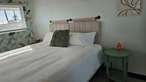 A neatly made bed with a headboard, a green side table with a salt lamp, and a window with blinds. - Day in Paradise - O'side Palms (Oceanside)