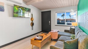 A modern living room with a wooden coffee table, a sofa, a TV mounted on the wall, and a window with a view of palm trees. - California Dreamin' - O'side Palms (Oceanside)