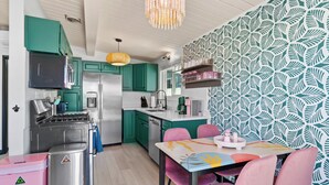 A kitchen with teal cabinets, a stainless steel refrigerator, a microwave, and a gas stove. There is a dining table with pink chairs and a colorful tablecloth. - Amazing Place by the Sea - O'side Palms (Oceanside)