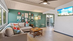 A living room with a ceiling fan, a large window, a green wall with framed pictures, a wooden coffee table, and a view of the outdoors. - Beach Meets Fabulous - O'side Palms (Oceanside)