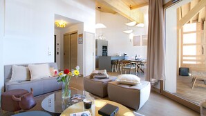 Ein modernes Wohnzimmer mit einer Couch, Hocker und Couchtisch, ein Essbereich mit Tisch und Stühlen sowie eine Küche im Hintergrund.