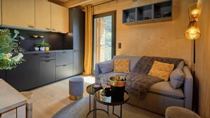 A modern living space with a grey sofa, a small round table, and a kitchen area with wooden cabinets and a window with curtains. - Chalet Sideflip - cosy stay for 4 with wonderful views - OVO Network (La Clusaz)