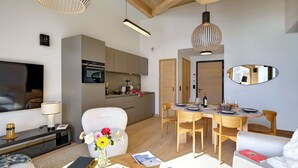 A modern living room with a TV, sofa, dining table, and chairs. There is a kitchen area with cabinets and appliances.