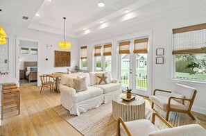 Amplia sala de estar con sofá blanco, sillas de madera, mesa de comedor y ventanas grandes con persianas.