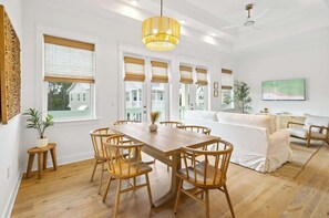 Un espace repas avec une table et des chaises en bois, un luminaire suspendu et une vue sur l’extérieur grâce à de grandes fenêtres.