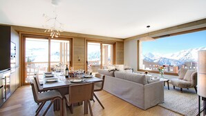 Un vaste espace de vie avec une grande fenêtre donnant sur un paysage montagneux, un coin repas avec une table en bois dressée pour quatre personnes, et un coin salon confortable avec un canapé et des fauteuils.