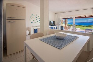 A kitchen with a white refrigerator, a dining table with a blue placemat, and a view of the sea through a window.