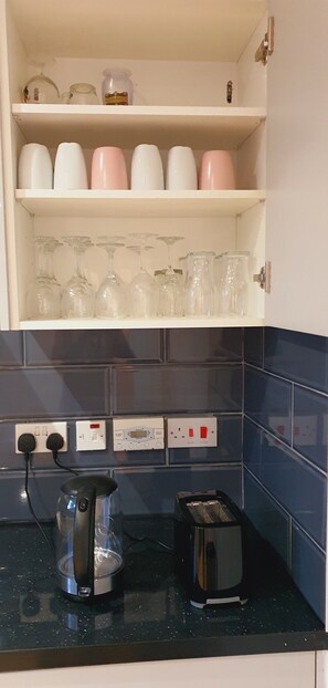 A kitchen with a countertop, a kettle, a toaster, and a shelf with glassware and jars. - Marahome (Watford)