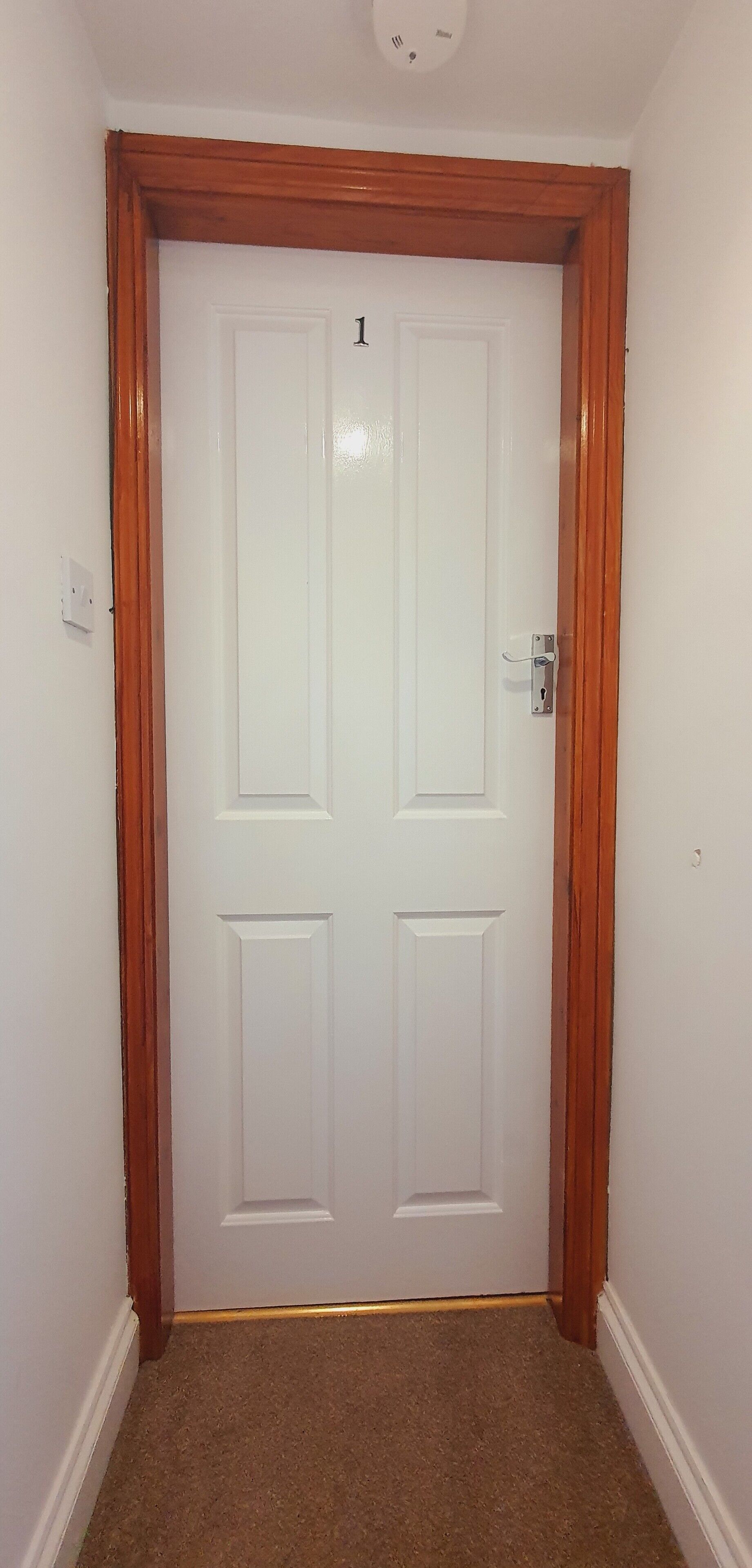 A white door with a number 1 above it, framed by wood, in a hallway with carpeted flooring.