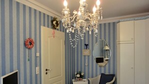 A room with striped blue and white walls, a white door, a white desk with a computer, a wicker chair with a blue cushion, a chandelier, and a mirror on the wall.