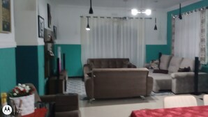 A living room with a brown sofa, a white armchair, and a dining table with a red tablecloth. There is a fireplace and a window with curtains. - Paraíso da Natureza (Ibiúna)