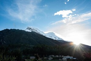 A mountainous landscape with a snow-capped peak and a sun setting or rising behind it. There are buildings in the foreground, likely a small settlement or village.