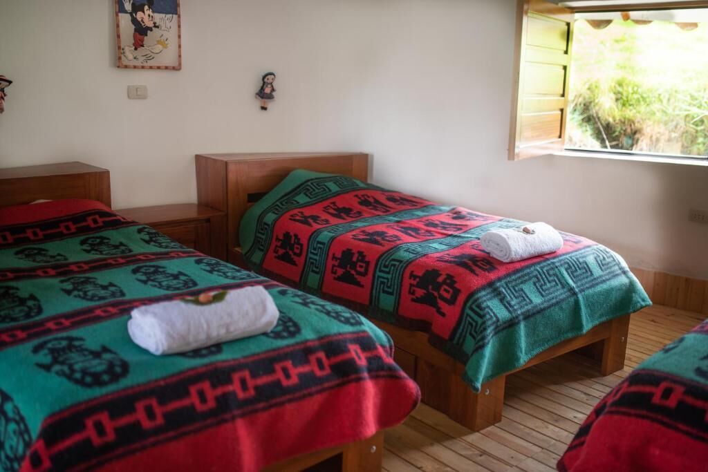 Quarto com duas camas de madeira, cada uma com um cobertor estampado e uma toalha enrolada. Há uma janela com vista para vegetação lá fora.