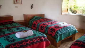 Quarto com duas camas de madeira, cada uma com um cobertor estampado e uma toalha enrolada. Há uma janela com vista para vegetação lá fora.