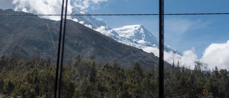A window view of a mountain with a snow-capped peak, a forested area, and some buildings in the foreground.