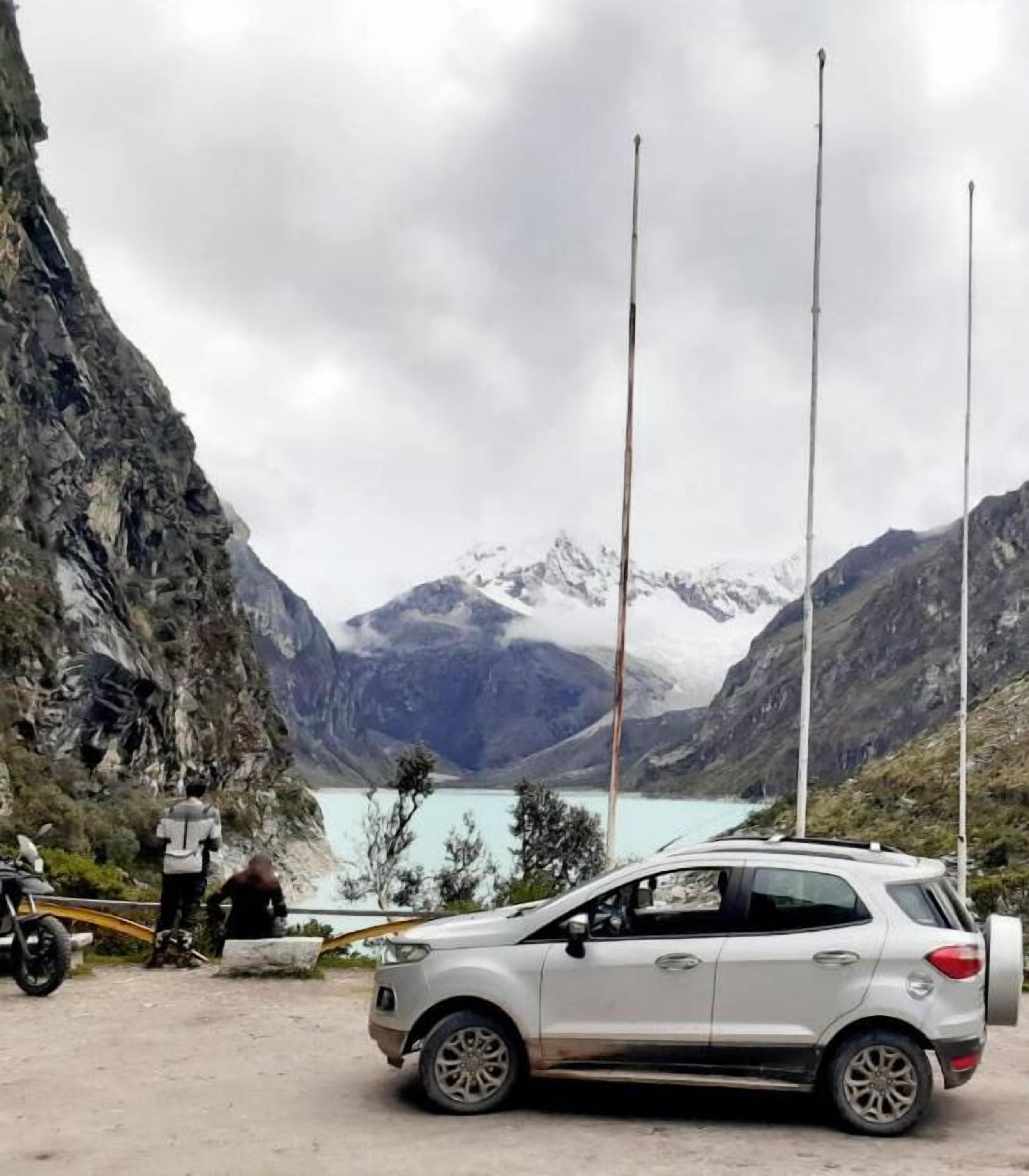 A white SUV parked near a rocky cliff with a lake and mountains in the background.