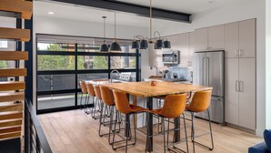 Een moderne keuken met een houten tafel, barkrukken, roestvrijstalen apparatuur en grote ramen.