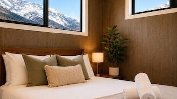 A bedroom with a bed, pillows, a bedside lamp, a plant, and a view of snow-capped mountains through two windows.