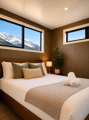 Une chambre à coucher avec un lit, des oreillers, une lampe de chevet, une plante et une vue sur des montagnes enneigées, visible à travers deux fenêtres.