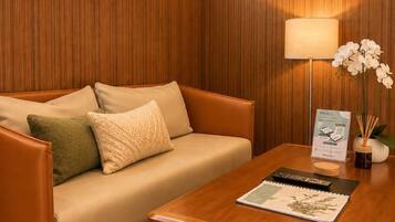 A room with a wooden paneled wall, a beige sofa with cushions, a wooden coffee table with a remote control and a magazine, and a lamp with an orchid.