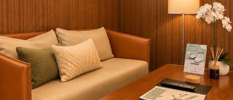 A room with a wooden paneled wall, a beige sofa with cushions, a wooden coffee table with a remote control and a magazine, and a lamp with an orchid.