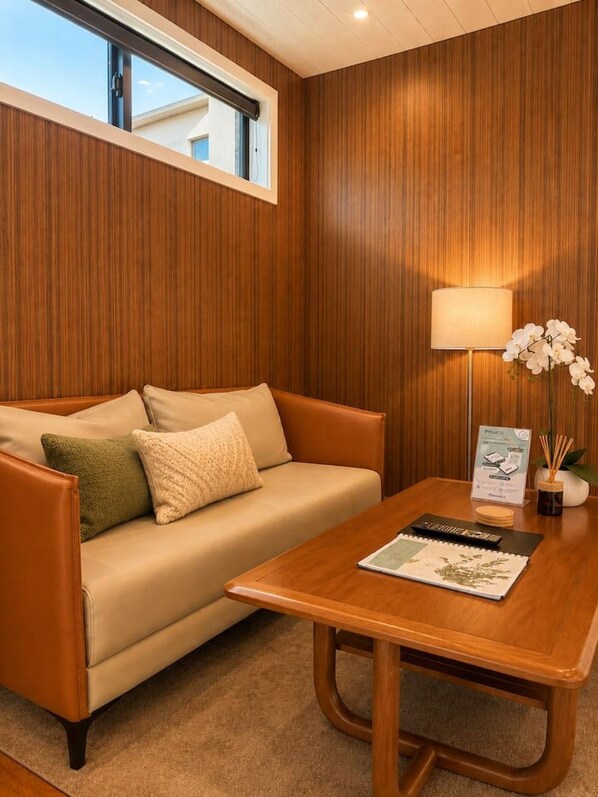 Une pièce avec un mur lambrissé en bois, un canapé beige avec des coussins, une table basse en bois sur laquelle se trouvent une télécommande et un magazine, et une lampe agrémentée d’une orchidée.