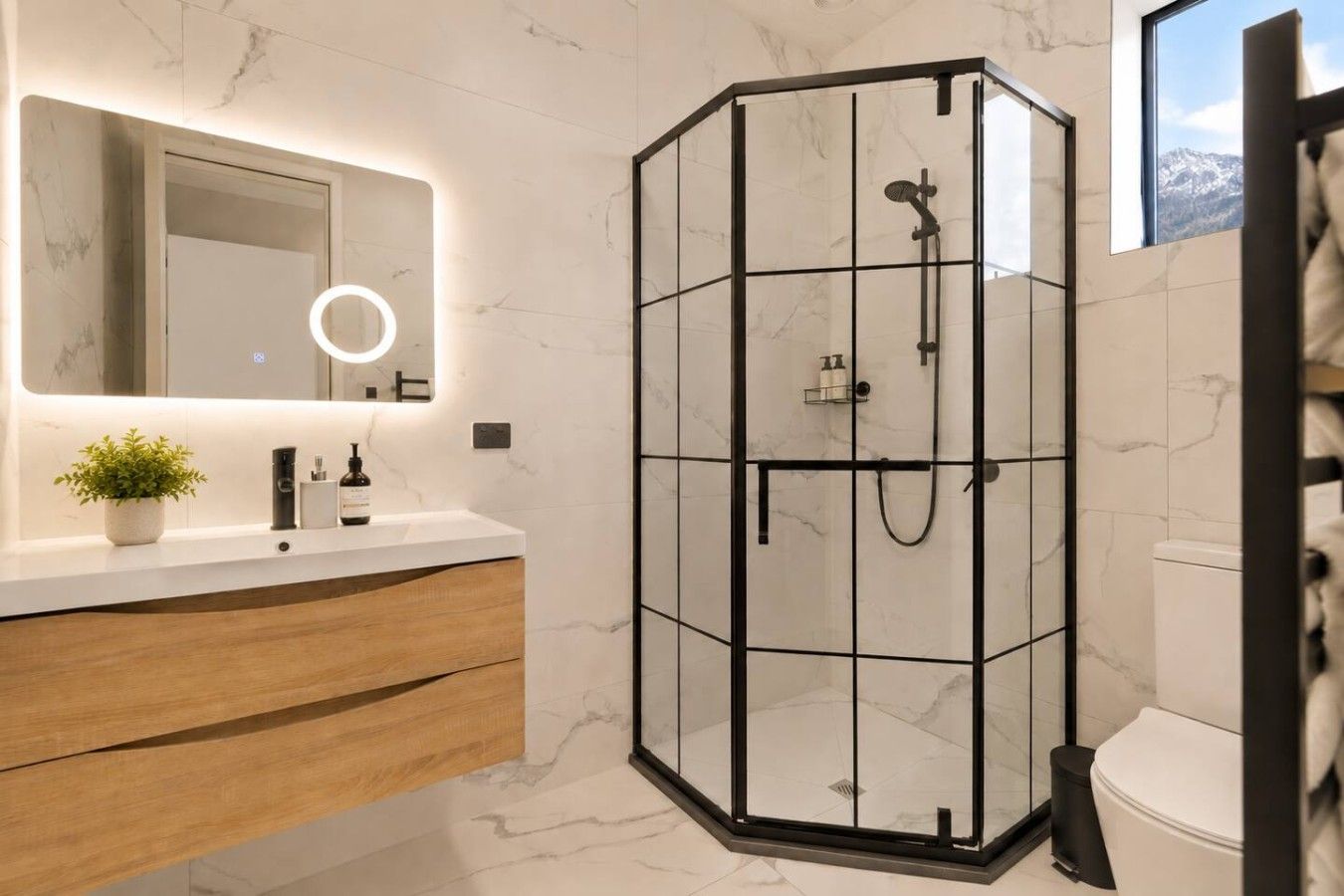 A modern bathroom with a glass-enclosed shower, a marble-tiled wall, a wooden vanity, and a large mirror with a ring light.