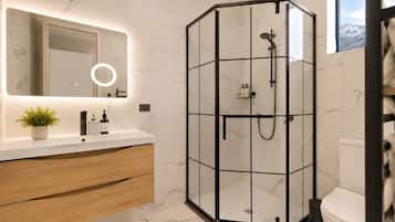 A modern bathroom with a glass-enclosed shower, a marble-tiled wall, a wooden vanity, and a large mirror with a ring light.