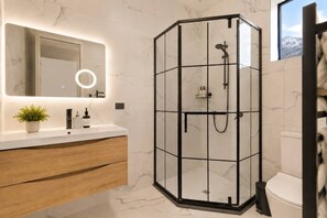 Une salle de bain moderne dotée d’une douche à parois de verre, d’un mur recouvert de carreaux de marbre, d’un meuble-lavabo en bois et d’un grand miroir avec un anneau lumineux.