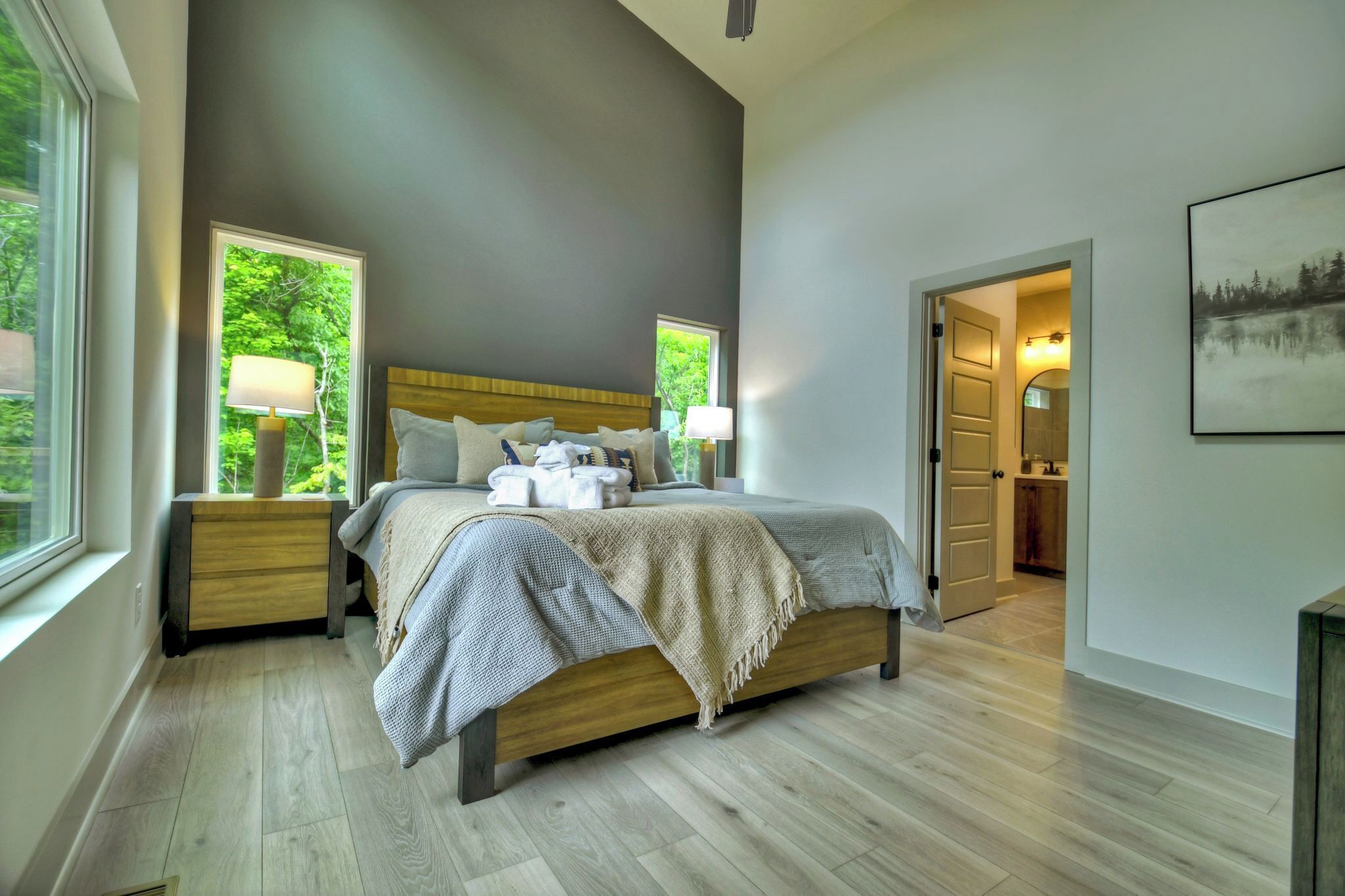 Une chambre moderne avec un grand lit, deux tables de chevet avec des lampes, une vue sur la verdure par une fenêtre et une porte donnant sur une salle de bain.