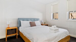 A neatly made bed with white linens, a blue headboard, and a small bedside table with a lamp and a plant.