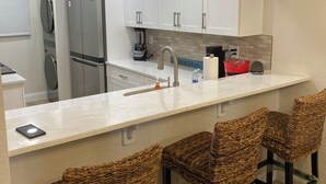 Una cocina moderna con armarios blancos, un refrigerador de acero inoxidable, una cafetera integrada y una zona de bar con sillas de mimbre.
