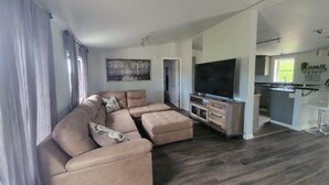 A modern living room with a sectional sofa, a flat-screen TV, and a kitchen area visible through an open doorway.