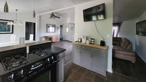 A modern kitchen with a gas stove, a built-in oven, and a dining area with a table and chairs. There is a ceiling fan, a flat-screen TV mounted on the wall, and a coffee maker on the counter.