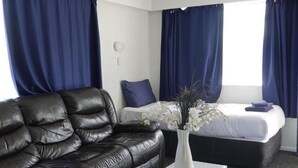 Una camera d'albergo con un divano in pelle nera, un vaso bianco con fiori, un letto con materasso bianco e tende blu, e un tavolino.