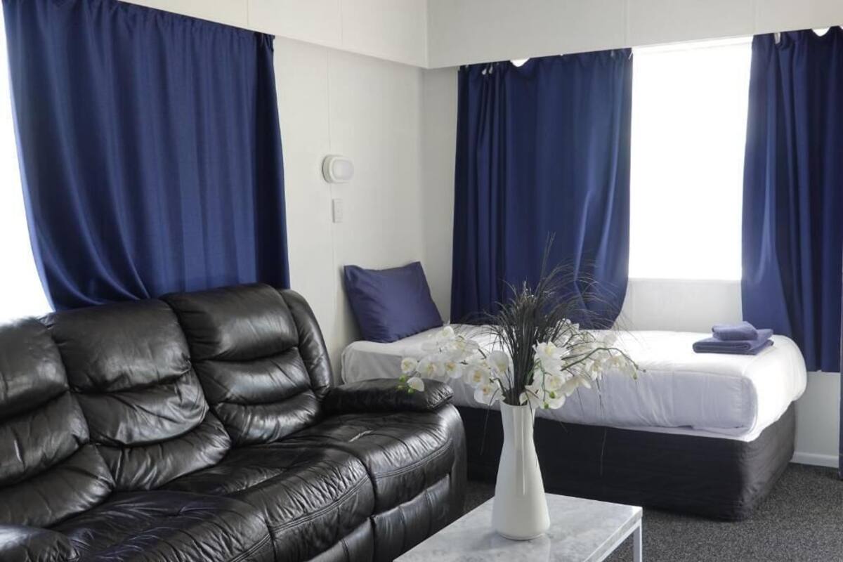 A hotel room with a black leather sofa, a white vase with flowers, a bed with a white mattress and blue curtains, and a small table.
