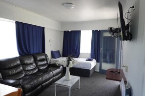 A modern hotel room with a black leather sofa, a bed with blue curtains, a small table with a vase of flowers, and a flat-screen TV mounted on the wall. - Ascot Lodge Motel  (Hamilton)